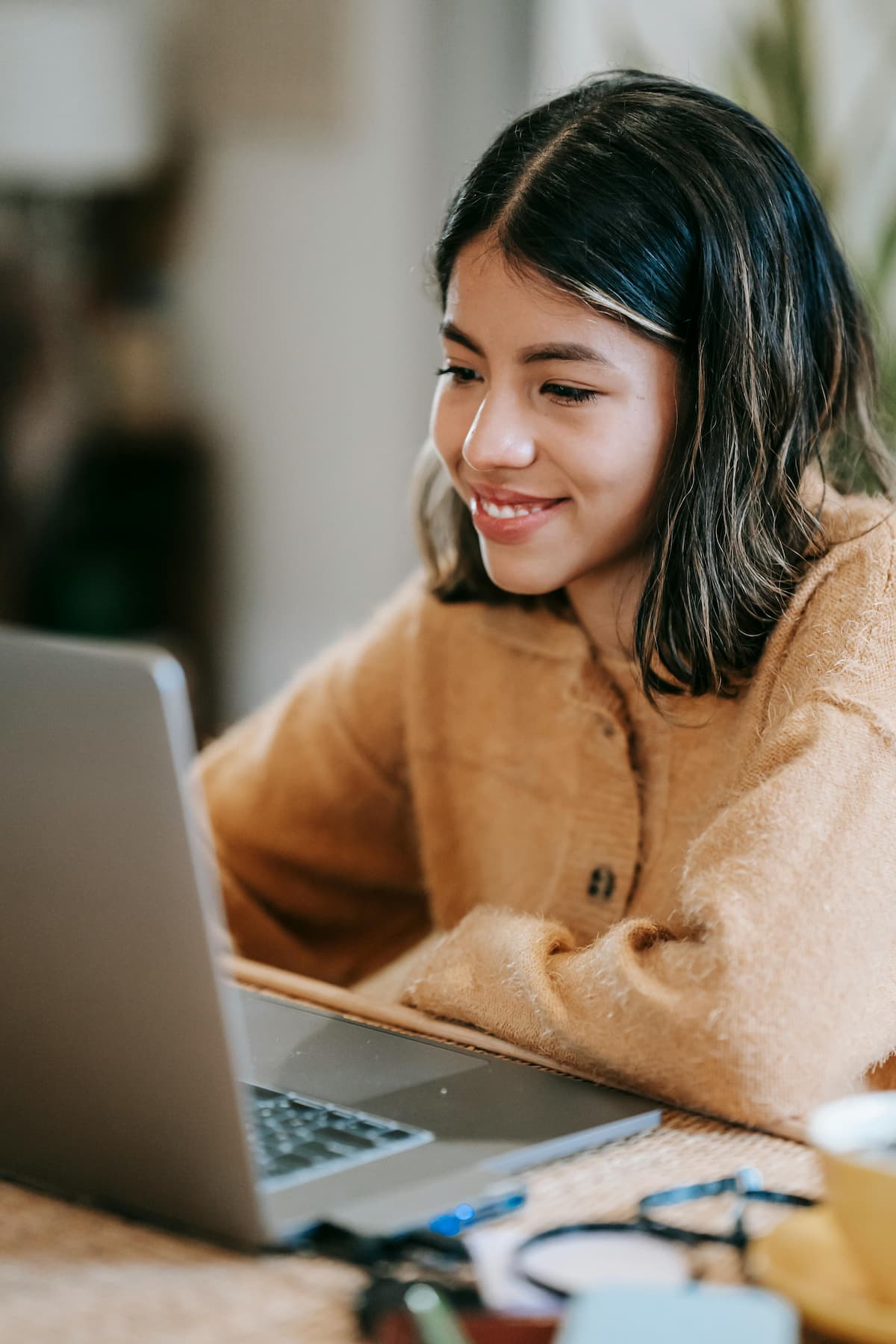 A young woman with dark hair and an orange sweater smiles while talking to a therapist on her laptop.